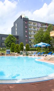 Hotel-Außenpool mit Sonnenliegen, Schirmen und dem Aparthotel am Rennsteig im Hintergrund bei blauem Sommerhimmel.