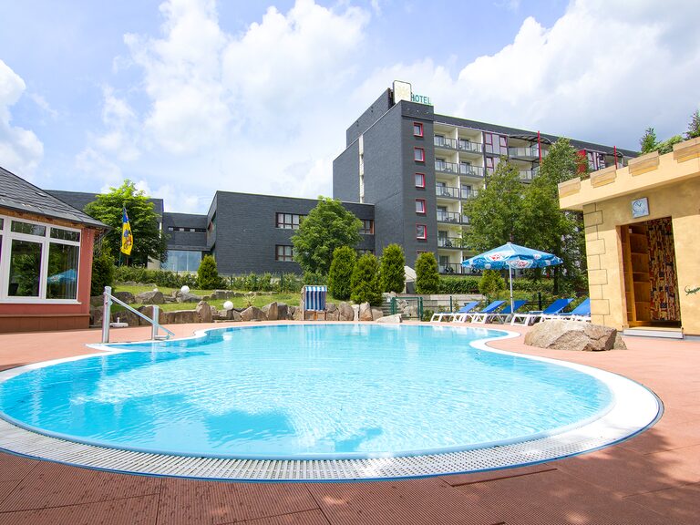 Hotel-Außenpool mit Sonnenliegen, Schirmen und dem Aparthotel am Rennsteig im Hintergrund bei blauem Sommerhimmel.