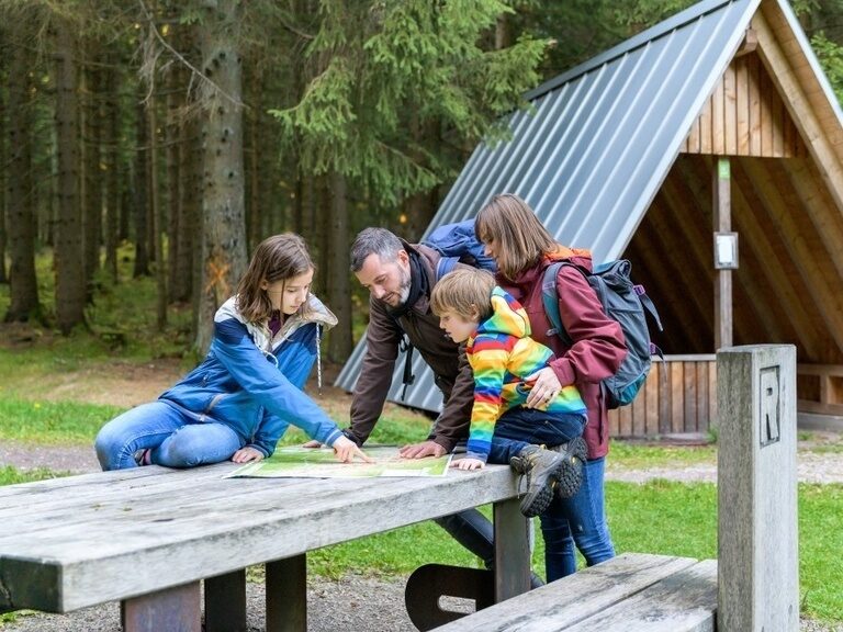 Familie bei einer Wanderung am Rennsteig vor einer Schutzhütte, sie studieren gemeinsam die Wanderkarte an einer Rennsteig-Bank