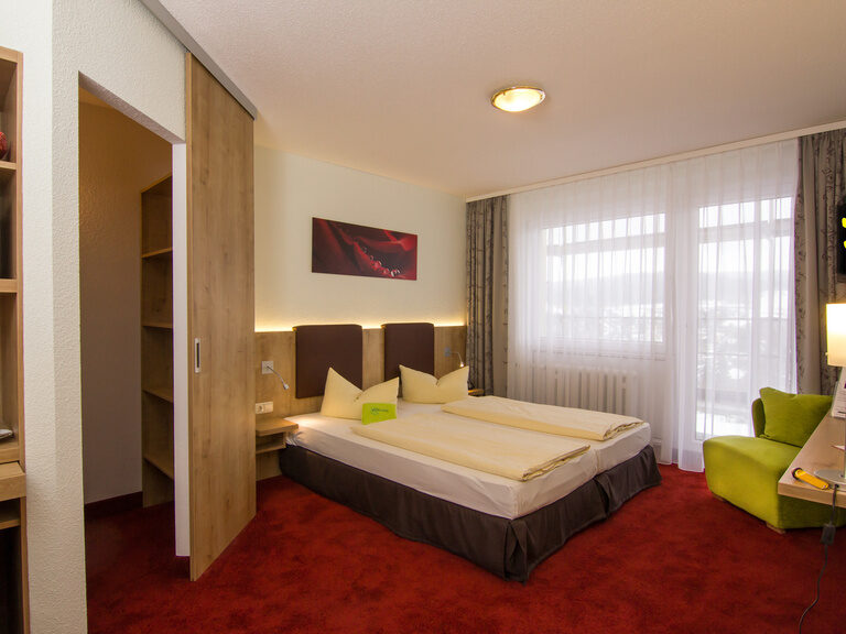 Doppelbett in einem Hotelzimmer mit Blick durch das Fenster in die umliegende Natur. Verweis zur Bildergalerie.