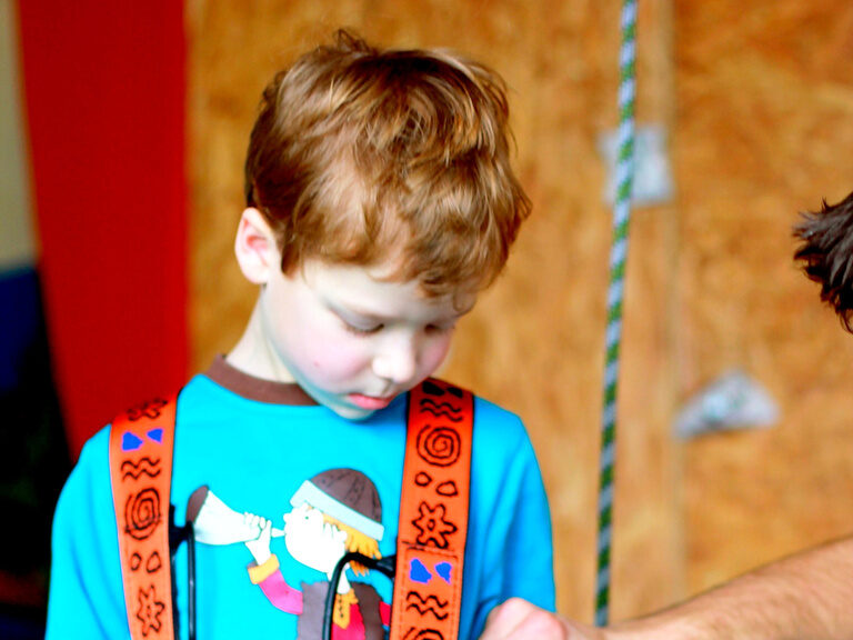 Sicher und abenteuerlustig: Ein kleines Mädchen in Matschanzug und Gummistiefeln klettert mutig über einen moosbewachsenen Baumstamm. Perfekt für einen Urlaub mit Enkeln und Kindern, die die Natur lieben.